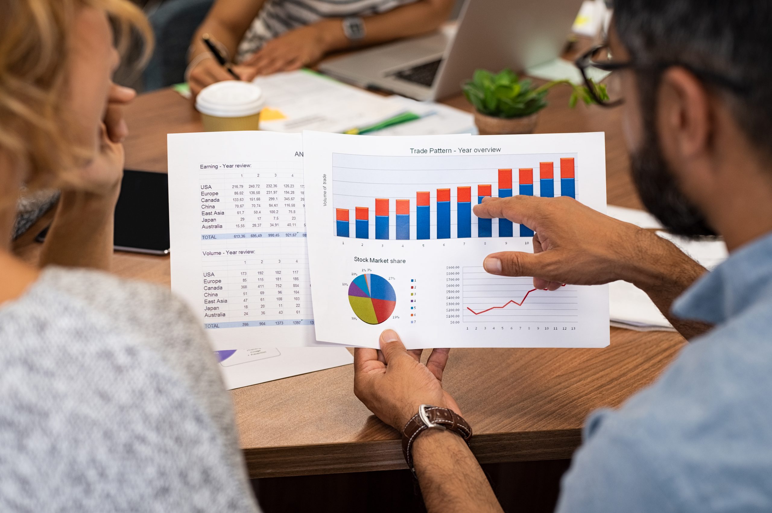 Mature latin businessman showing stock market bar graph to businesswoman. Financial advising team discussing trading patterns of year. Rear view of casual businesspeople holding charts studying year trend together.