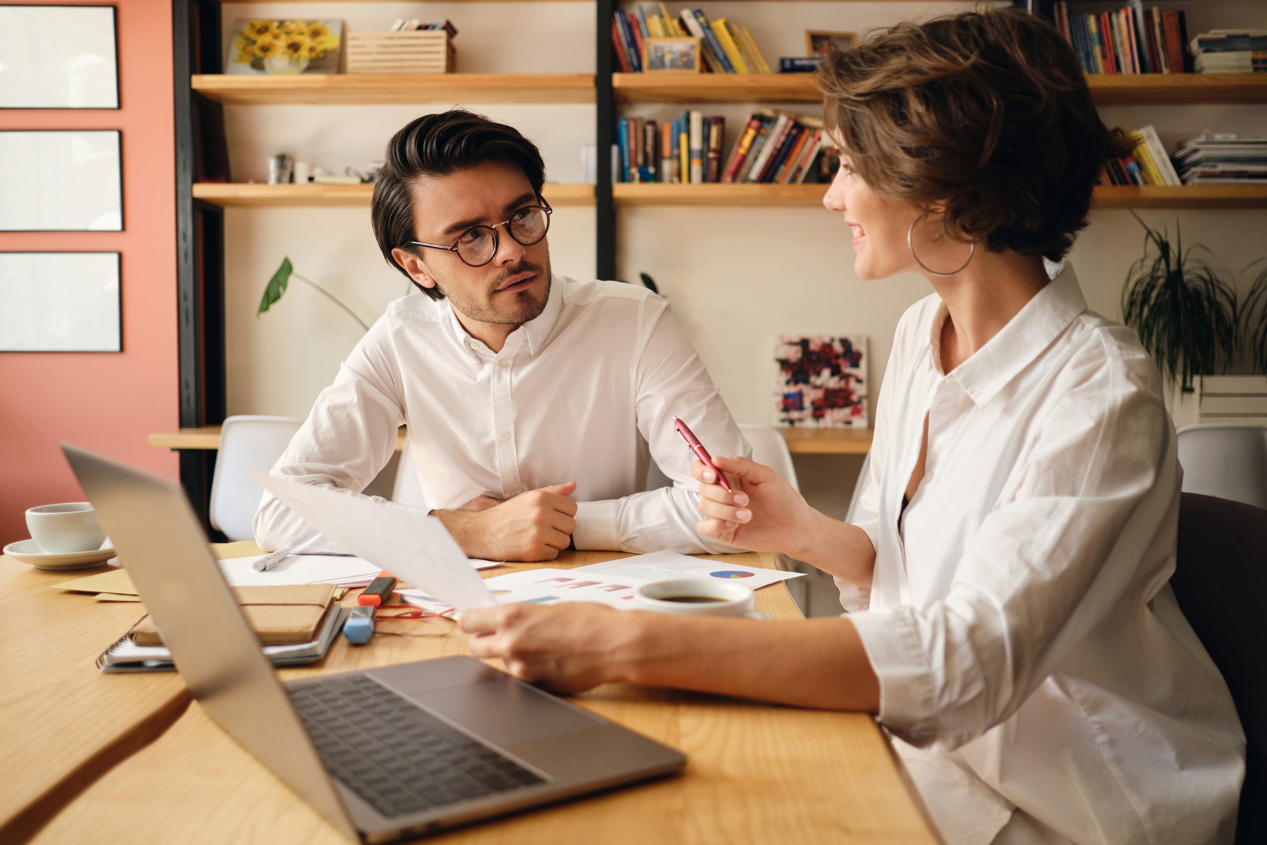 Young attractive business colleagues thoughtfully discussing new project sitting at the table with laptop and papers in modern office